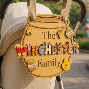 May include: A yellow honey pot-shaped sign hanging from a stroller handle. The sign features the text "The WINCHESTER Family" with colorful character-themed letters, a small beehive, a honey dipper, two honey pots, and a Winnie the Pooh face icon.