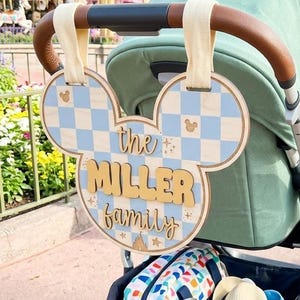May include: A wooden stroller sign shaped like a mouse head with a light blue and white checkered pattern. The sign features the text 'the MILLER family' in raised, tan-colored lettering, attached to the stroller handle with two cream fabric straps.