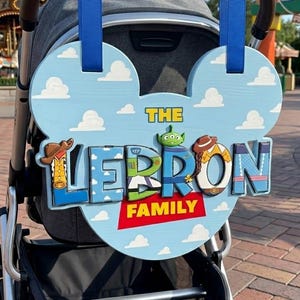 May include: A light blue, Mickey-shaped wooden sign with white clouds, hanging from a stroller by two blue ribbons. The sign features the text THE LEBRON FAMILY in colorful, character-themed letters, including a green alien and cowboy motifs.