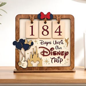 May include: A wooden countdown sign featuring three hanging rectangular tiles displaying the number 184. The sign includes a red bow, a gold castle silhouette, dark blue balloons, and the text "Days Until Our Disney Trip" in maroon.