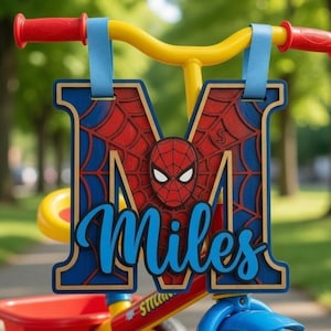 May include: A personalised wooden name sign featuring the letter M with a red and blue spiderweb pattern and a Spider-Man mask graphic. The name Miles is written in blue script across the front. The sign hangs from the yellow handlebars of a tricycle.