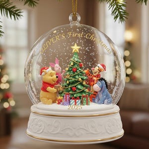 Puede incluir: Un globo de nieve de cristal transparente con un lazo dorado para colgar. En el interior, los personajes de Winnie the Pooh rodean un árbol de Navidad. La base es blanca con adornos dorados, y el texto "OLIVER'S First Christmas" está en el globo. Se muestra el año 2025.