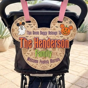 May include: A wooden stroller sign shaped like mouse ears with pink straps, featuring a ghost, a pumpkin, and spiderweb patterns. The text reads: This Doom Buggy Belongs To The Henderson Family Welcome Foolish Mortals in orange and green.