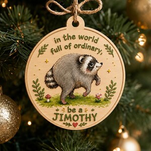 Puede incluir: Un adorno navide&ntilde;o redondo de madera con una ilustraci&oacute;n impresa de un mapache sobre un parche de hierba verde. El texto dice, in the world full of ordinary, be a JIMOTHY. El adorno est&aacute; suspendido por un lazo de cuerda marr&oacute;n en la parte superior.