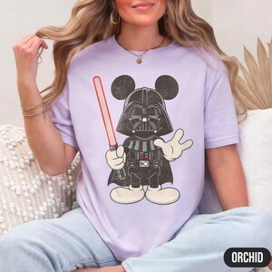 Pu&ograve; includere: Una maglietta lilla con un'illustrazione a cartoni animati di Darth Vader con le orecchie di Topolino, che tiene una spada laser rossa. Il design &egrave; in nero, bianco e grigio. La parola "ORCHID" &egrave; stampata in un ovale nero in basso a destra.