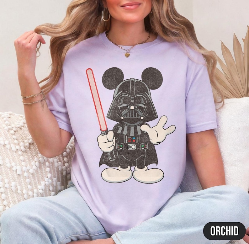 Pu&ograve; includere: Una maglietta lilla con un'illustrazione a cartoni animati di Darth Vader con le orecchie di Topolino, che tiene una spada laser rossa. Il design &egrave; in nero, bianco e grigio. La parola "ORCHID" &egrave; stampata in un ovale nero in basso a destra.
