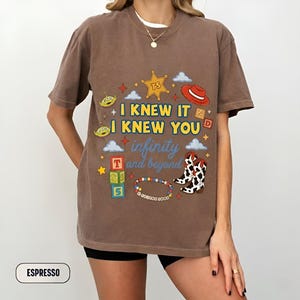 Puede incluir: Una camiseta de manga corta color marr&oacute;n espresso con un estampado gr&aacute;fico que incluye el texto I KNEW IT I KNEW YOU infinity and beyond. El dise&ntilde;o presenta una estrella de sheriff amarilla, un sombrero de vaquero rojo, aliens y botas.