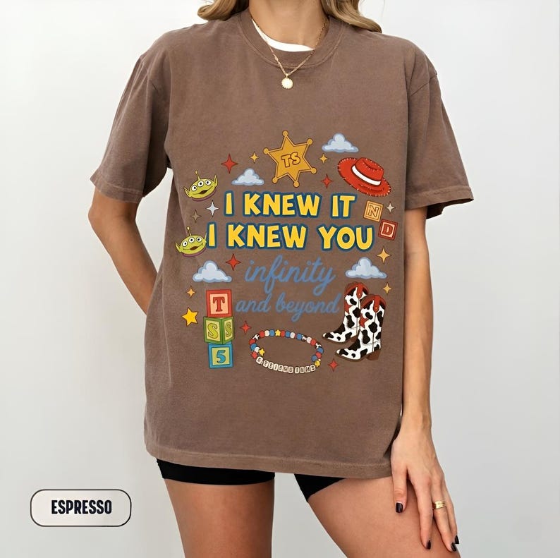 Puede incluir: Una camiseta de manga corta color marr&oacute;n espresso con un estampado gr&aacute;fico que incluye el texto I KNEW IT I KNEW YOU infinity and beyond. El dise&ntilde;o presenta una estrella de sheriff amarilla, un sombrero de vaquero rojo, aliens y botas.