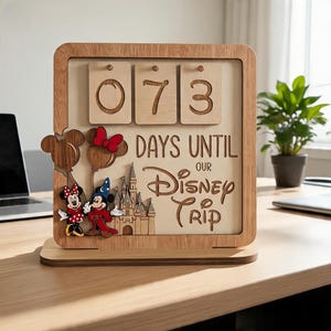 Puede incluir: Un calendario de cuenta regresiva de madera con tres tarjetas numéricas abatibles que muestran 073. El texto dice DAYS UNTIL OUR DISNEY TRIP. Incluye figuras de madera cortadas con láser de dos ratones, globos y un castillo.