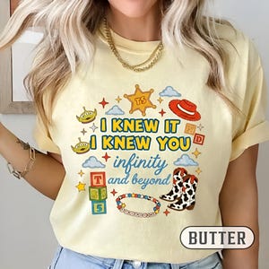 Puede incluir: Una camiseta de color amarillo p&aacute;lido con un estampado gr&aacute;fico que incluye el texto I KNEW IT I KNEW YOU infinity and beyond. El dise&ntilde;o presenta ilustraciones de dos alien&iacute;genas verdes, una estrella de sheriff, un sombrero rojo y botas.