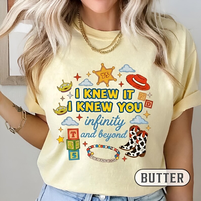 Puede incluir: Una camiseta de color amarillo p&aacute;lido con un estampado gr&aacute;fico que incluye el texto I KNEW IT I KNEW YOU infinity and beyond. El dise&ntilde;o presenta ilustraciones de dos alien&iacute;genas verdes, una estrella de sheriff, un sombrero rojo y botas.
