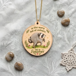 Puede incluir: Un colgante redondo de madera en una cadena dorada con una ilustraci&oacute;n impresa de un mapache en un &aacute;rea de hierba con setas. El texto dice: in the world full of ordinary, be a JIMOTHY. El dise&ntilde;o incluye hojas verdes y un peque&ntilde;o coraz&oacute;n.