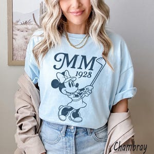 Puede incluir: Camiseta azul claro con un gr&aacute;fico en blanco y negro de Minnie Mouse jugando al golf y el texto "MM 1928". Esta camiseta informal est&aacute; hecha de una tela suave, perfecta para el uso diario.