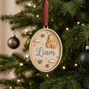 May include: An oval-shaped wooden Christmas ornament featuring a laser-cut Winnie the Pooh character holding a honey pot, small bees, and clouds. The name Liam is printed in blue script in the center. It hangs from a dark red ribbon.