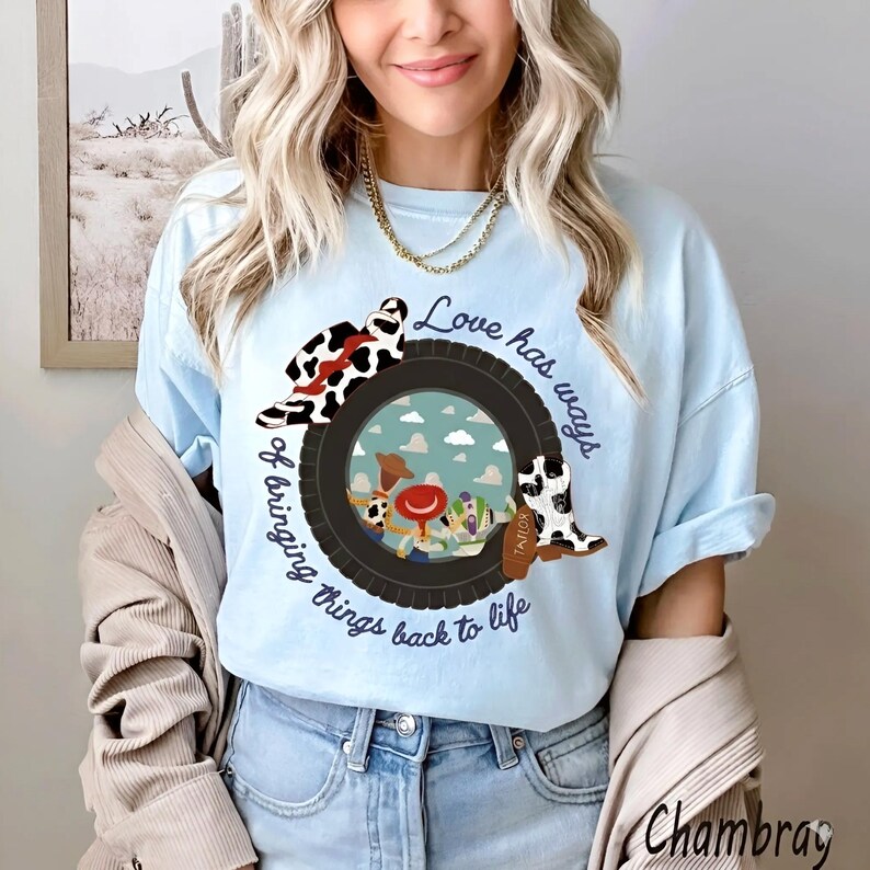 May include: A light blue t-shirt featuring a graphic of a black tire surrounding a sky-blue background with cartoon characters. The design includes a cow-print hat, a cow-print boot, and the text "Love has ways of bringing things back to life."