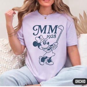 Puede incluir: Camiseta de color lila claro con un gr&aacute;fico azul marino de Minnie Mouse jugando al golf, con el texto "MM 1928". La camiseta tiene cuello redondo y mangas cortas. La palabra "ORCHID" est&aacute; impresa en un rect&aacute;ngulo negro en la parte inferior.