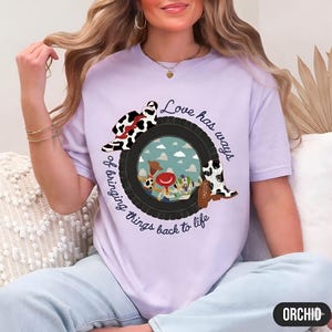 May include: A light purple t-shirt featuring a graphic of a black tire framing a sky scene with cartoon characters. The design includes a cow-print cowboy hat, a cow-print boot with the name Taylor, and the text Love has ways of bringing things back to life.