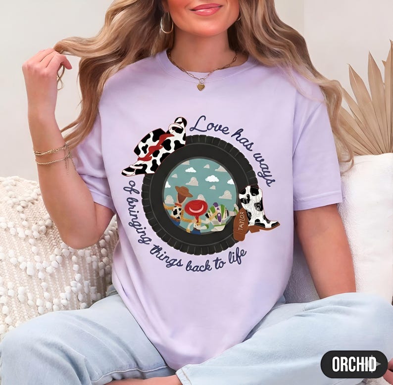 May include: A light purple t-shirt featuring a graphic of a black tire framing a sky scene with cartoon characters. The design includes a cow-print cowboy hat, a cow-print boot with the name Taylor, and the text Love has ways of bringing things back to life.