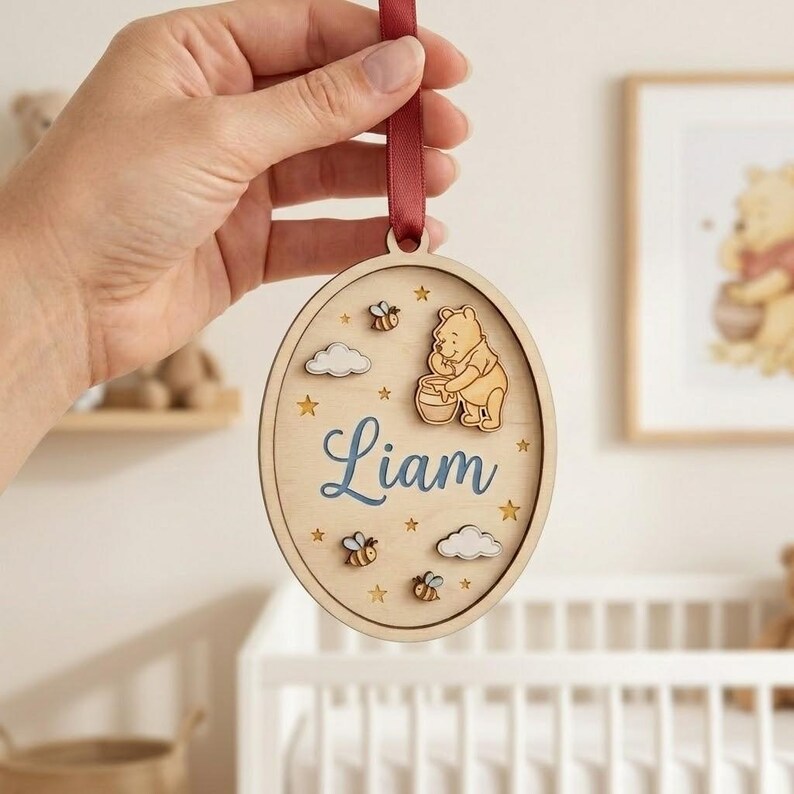 May include: An oval wooden hanging ornament featuring a laser-cut Winnie the Pooh character holding a honey pot, the name Liam in blue script, small bees, clouds, and star cutouts. The ornament is suspended by a dark red satin ribbon.