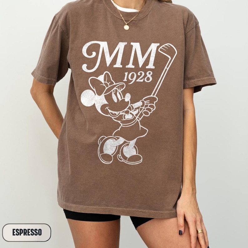 Puede incluir: Camiseta color espresso con un gr&aacute;fico blanco de un personaje de dibujos animados jugando al golf, con el texto "MM 1928". La camiseta tiene mangas cortas y cuello redondo. La palabra "ESPRESSO" est&aacute; en un rect&aacute;ngulo blanco en la parte inferior izquierda.