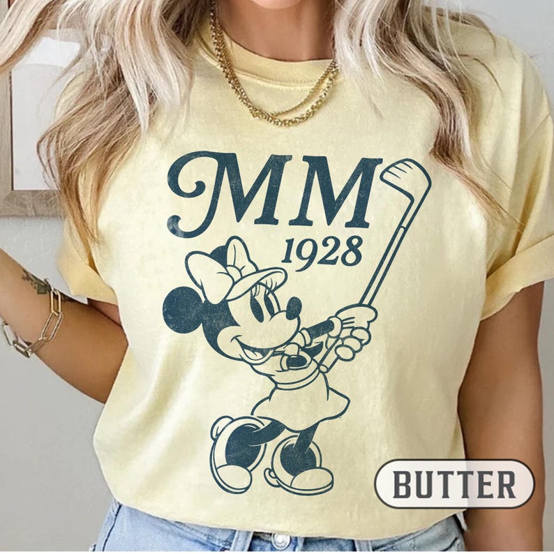 Puede incluir: Camiseta de color mantequilla con un gr&aacute;fico azul marino de Minnie Mouse jugando al golf, con el texto "MM 1928". La palabra "BUTTER" est&aacute; impresa en un recuadro en la parte inferior. La camiseta tiene mangas cortas.