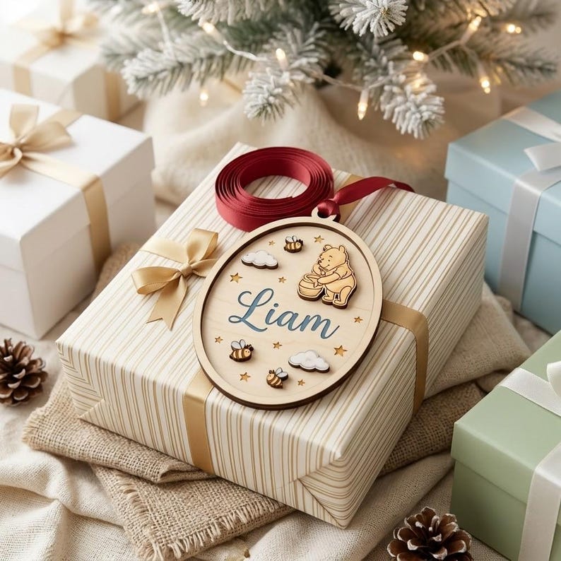 May include: An oval wooden ornament featuring a laser-cut Winnie the Pooh character holding a honey pot, surrounded by small bees, clouds, and stars. The name Liam is printed in blue script in the center. A dark red ribbon is attached to the top.