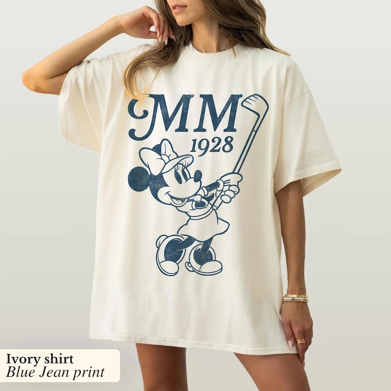 Puede incluir: Camiseta de color marfil con un gr&aacute;fico azul marino de Minnie Mouse jugando al golf, con el texto "MM 1928". La camiseta tiene mangas cortas y un corte holgado. El texto "Ivory shirt Blue Jean print" est&aacute; en la parte inferior de la imagen.
