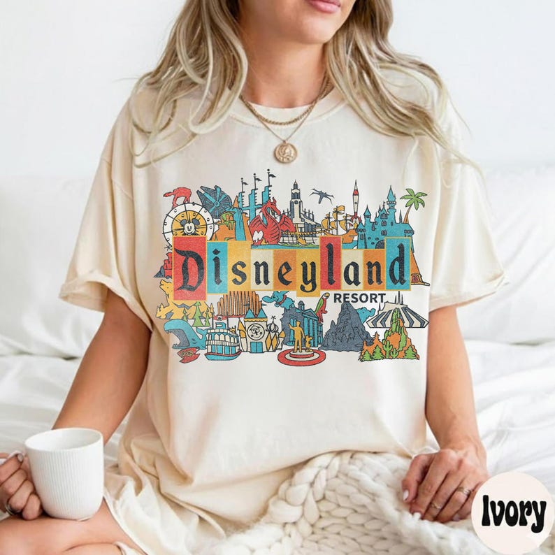 K&ouml;nnte beinhalten: Cremefarbenes T-Shirt mit einem farbenfrohen Disneyland Resort-Motiv. Die Grafik enth&auml;lt das Disneyland-Logo und verschiedene Parksymbole. Das Shirt hat kurze &Auml;rmel und eine lockere Passform.