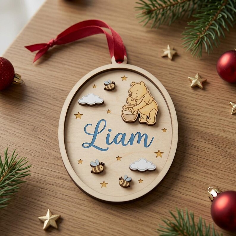 May include: An oval wooden Christmas ornament featuring a laser-cut Winnie the Pooh character holding a honey pot, three small bees, two clouds, and several star cutouts. The name Liam is printed in blue script in the center with a red ribbon.