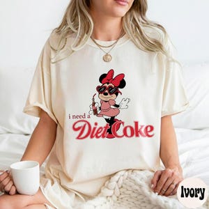 May include: Cream-coloured t-shirt with a Minnie Mouse graphic wearing sunglasses, a pink dress, and holding a Diet Coke. The text "i need a Diet Coke" is printed in red script. The shirt has a relaxed, oversized fit.