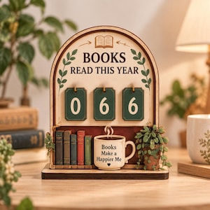 May include: An arched wooden book tracker sign with the text BOOKS READ THIS YEAR, featuring three green flip-style number cards currently set to 066. Below are miniature book models, a coffee mug labeled Books Make a Happier Me, and a potted plant.