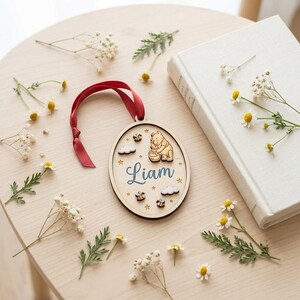 May include: An oval wooden ornament featuring a laser-cut Winnie the Pooh character, a honey pot, small bees, clouds, and stars. The name Liam is printed in blue script in the center. A dark red ribbon is attached to the top for hanging.