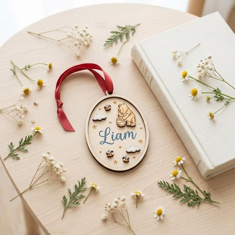 May include: An oval wooden ornament featuring a laser-cut Winnie the Pooh character, a honey pot, small bees, clouds, and stars. The name Liam is printed in blue script in the center. A dark red ribbon is attached to the top for hanging.