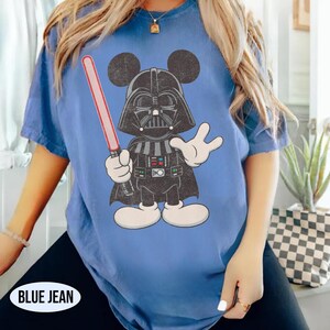 Pu&ograve; includere: T-shirt blu jeans con un personaggio dei cartoni animati ibrido di Darth Vader e Topolino. Il personaggio tiene una spada laser rossa e indossa un casco e un mantello neri. La maglietta ha il testo "BLUE JEAN" in basso.