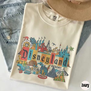 K&ouml;nnte beinhalten: Elfenbeinfarbenes T-Shirt mit einem farbenfrohen Disneyland Resort-Grafik. Das Design zeigt ikonische Parkbilder und das Wort "Disneyland" in einer kr&auml;ftigen, mehrfarbigen Schrift. Das Shirt liegt gefaltet auf einer Jeans und einer Halskette.