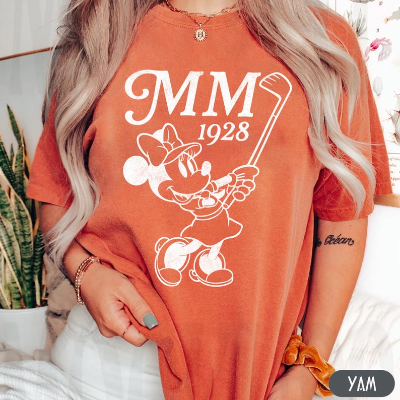 Puede incluir: Camiseta naranja quemado con un gr&aacute;fico blanco de Minnie Mouse jugando al golf, con el texto "MM 1928". El dise&ntilde;o incluye a Minnie Mouse con un atuendo de golf, sosteniendo un palo de golf. La camiseta es de estilo casual.