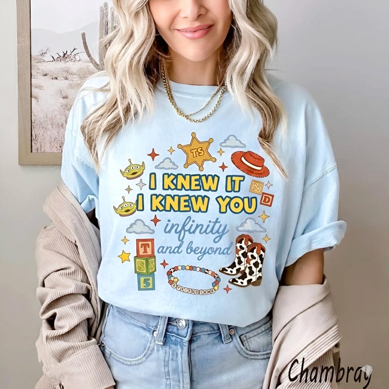 Puede incluir: Una camiseta azul claro con un estampado gr&aacute;fico que incluye el texto I KNEW IT I KNEW YOU infinity and beyond. El dise&ntilde;o presenta ilustraciones de una estrella de sheriff, alien&iacute;genas, un sombrero de vaquero, bloques, botas y una pulsera.