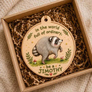 Puede incluir: Un adorno redondo de madera con un mapache pintado sobre hierba verde. El texto dice: "in the world full of ordinary, be a JIMOTHY". El dise&ntilde;o incluye peque&ntilde;as ramas verdes, diminutas estrellas amarillas, una seta roja y un peque&ntilde;o coraz&oacute;n rojo.
