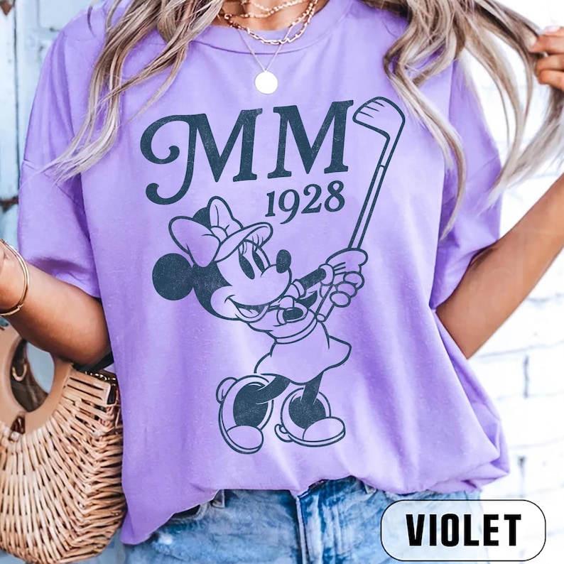Puede incluir: Camiseta violeta con un dise&ntilde;o de Minnie Mouse jugando al golf, con el texto "MM 1928" en gris oscuro. La camiseta tiene mangas cortas y cuello redondo. La palabra "VIOLET" est&aacute; impresa en la parte inferior.