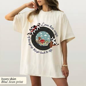 May include: An ivory t-shirt featuring a graphic print of a black tire surrounding a sky scene with cartoon characters. The design includes a cow-print hat, a cow-print boot, and the text "Love has ways of bringing things back to life."