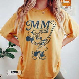 Puede incluir: Camiseta color mostaza con un gr&aacute;fico azul marino de Minnie Mouse jugando al golf, con el texto "MM 1928". La camiseta tiene mangas cortas y un corte holgado. La palabra "Mostaza" est&aacute; en un recuadro blanco en la parte inferior.