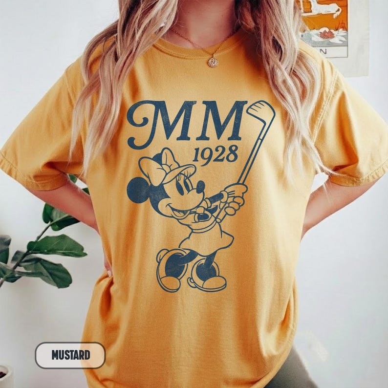 Puede incluir: Camiseta color mostaza con un gr&aacute;fico azul marino de Minnie Mouse jugando al golf, con el texto "MM 1928". La camiseta tiene mangas cortas y un corte holgado. La palabra "Mostaza" est&aacute; en un recuadro blanco en la parte inferior.