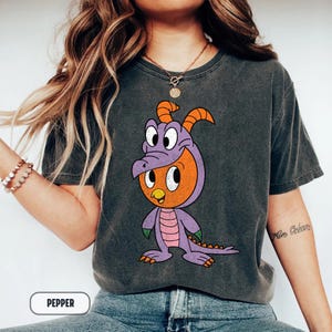 Puede incluir: Camiseta gris oscuro con un personaje de dibujos animados con un disfraz de dragón morado y naranja. El personaje tiene grandes ojos blancos y está de pie. La palabra "PEPPER" está impresa en la parte inferior de la imagen.