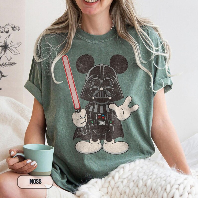 Pu&ograve; includere: T-shirt verde muschio con un personaggio dei cartoni animati di Darth Vader con orecchie di Topolino, che tiene una spada laser rossa. Il personaggio indossa un elmo e un mantello neri. La maglietta ha un aspetto vintage.