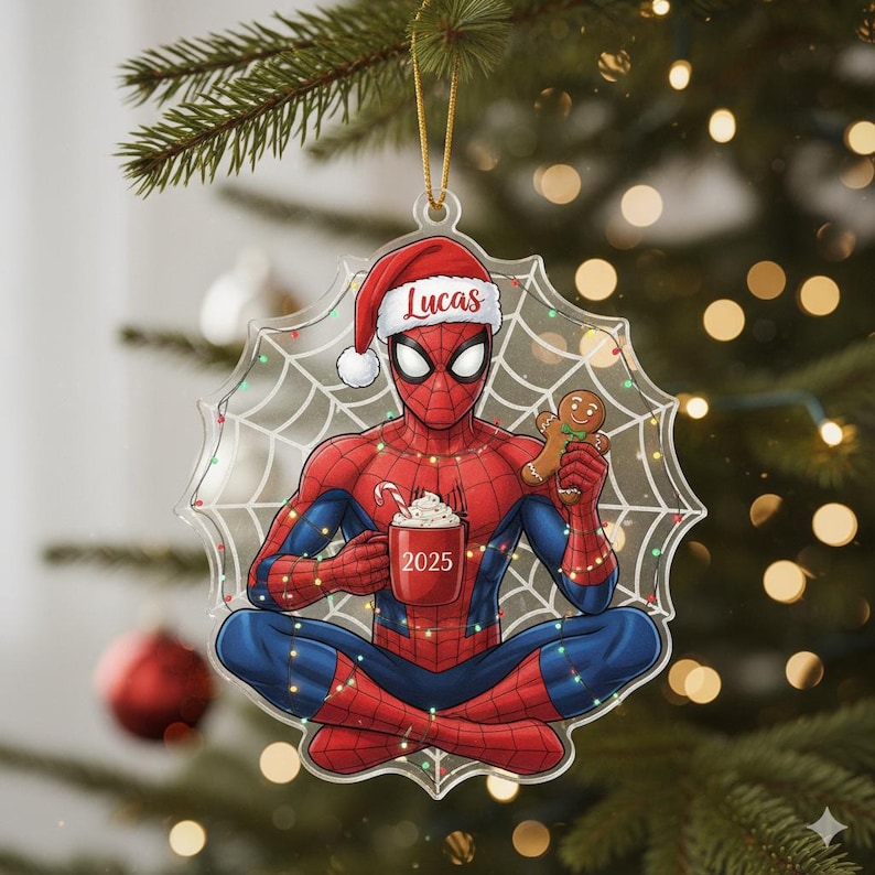May include: A clear acrylic Christmas ornament featuring Spider-Man in a Santa hat, holding a mug and gingerbread man. The ornament is shaped like a spider web and has the name "Lucas" on the hat and the year "2025" on the mug.