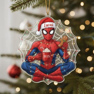 May include: Spider-Man Christmas ornament with the name "Lucas" on a Santa hat. The ornament features Spider-Man holding a mug with "2025" on it and a gingerbread man. The ornament is surrounded by a spider web design.
