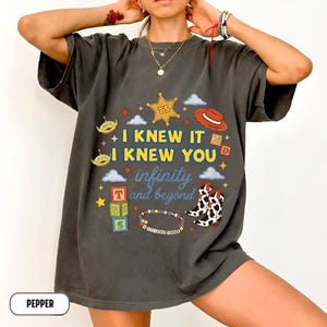 Puede incluir: Una camiseta gris carb&oacute;n de manga corta con un estampado gr&aacute;fico que incluye el texto I KNEW IT I KNEW YOU infinity and beyond. El dise&ntilde;o presenta una estrella de sheriff, un sombrero de vaquero, alien&iacute;genas, bloques y botas.
