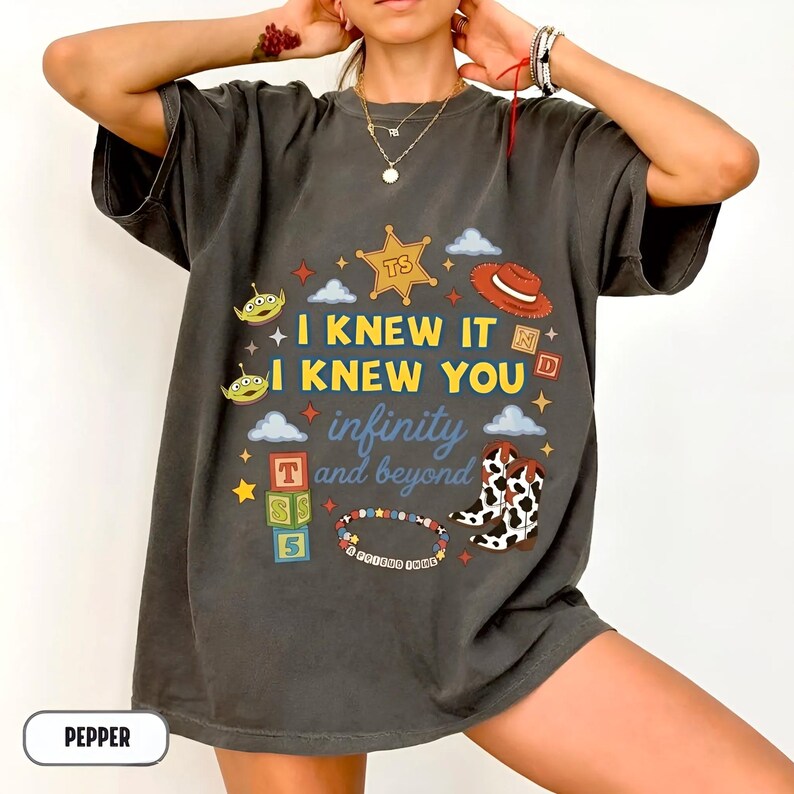 Puede incluir: Una camiseta gris carb&oacute;n de manga corta con un estampado gr&aacute;fico que incluye el texto I KNEW IT I KNEW YOU infinity and beyond. El dise&ntilde;o presenta una estrella de sheriff, un sombrero de vaquero, alien&iacute;genas, bloques y botas.
