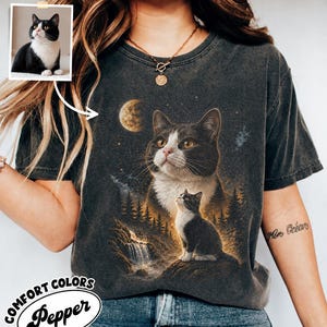 May include: A charcoal gray t-shirt featuring a graphic print of a black and white cat looking up at a crescent moon in a starry forest landscape. A smaller inset photo shows the same black and white cat. The shirt has a vintage wash finish.