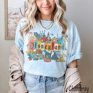 K&ouml;nnte beinhalten: Hellblaues T-Shirt mit einem farbenfrohen Disneyland Resort-Motiv. Das Design zeigt das Disneyland-Logo in Regenbogenfarben, umgeben von ikonischen Parkbildern. Das Shirt ist ein bequemer, l&auml;ssiger Stil.